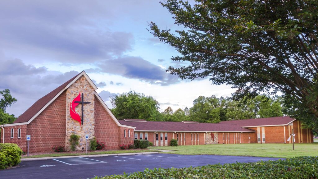 Wesley United Methodist Church is a brick building with a black cross and red flame emblem on the tallest wall. A tree is visible in the right side of the image.
