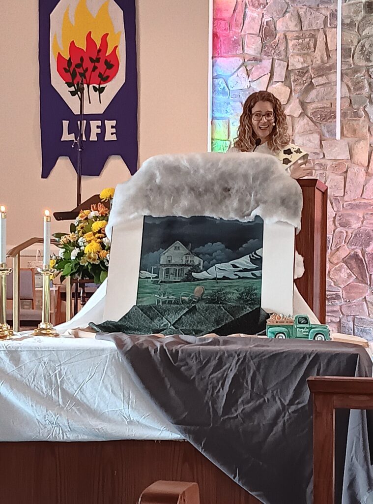 Pastor Kerry at the pulpit on "Storm Sunday" during our worship series on Creation. The altar has a print of a storm approaching a farmhouse. Above the print is grey batting to resemble clouds, and on the altar are gray fabric and a small green farm truck that matches the style of the painting.